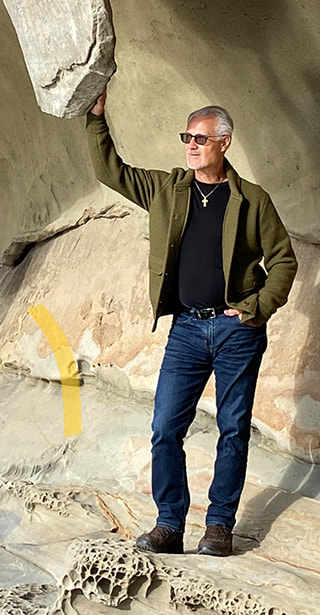 David Moss standing on some sandstone rocks in a green jacket, jeans and sunglasses on a sunny day